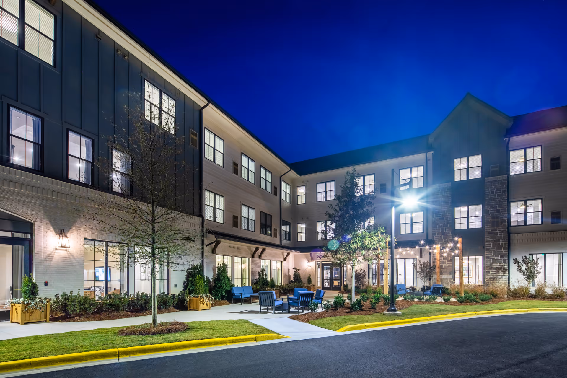 Exterior view of a modern three-story senior living facility at night with well-lit windows, outdoor seating areas with blue chairs, landscaped greenery, and a street lamp illuminating the entrance area.