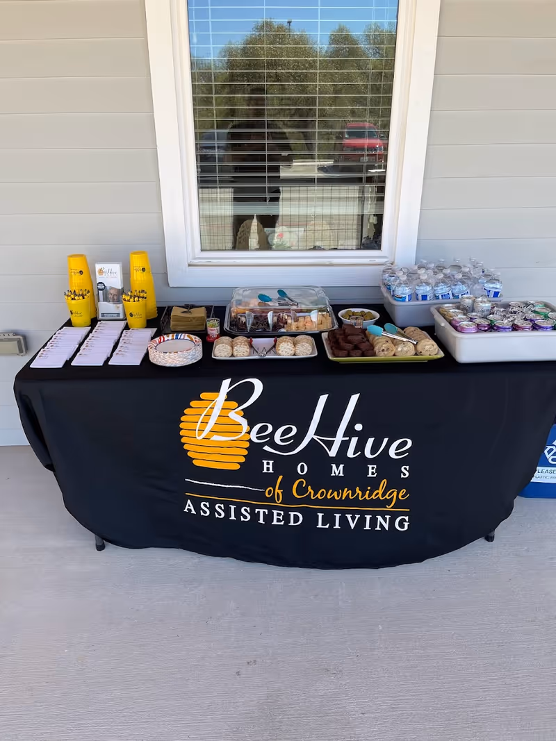A table covered with a black tablecloth displaying the logo and name 'BeeHive Homes of Crownridge Assisted Living' set up outdoors against a building wall with a window. The table holds various snacks including crackers, brownies, cookies, bottled water, and canned beverages, along with yellow cups and informational brochures.