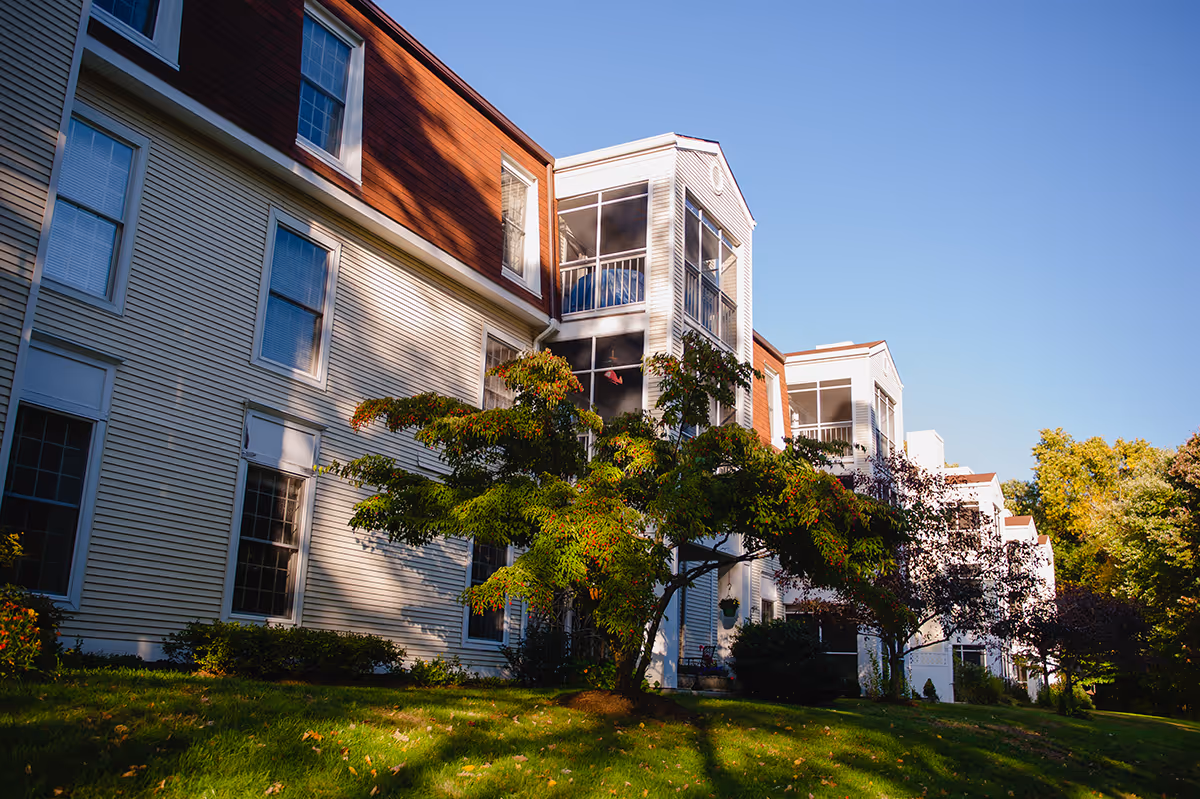 Exterior view of a multi-story residential building with screened balconies, windows, and trees on a grassy lawn.