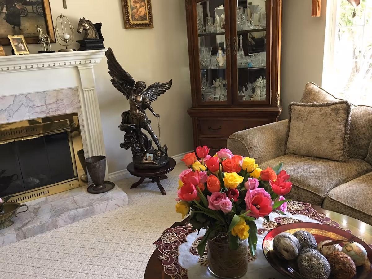 A cozy living room with a beige patterned sofa and matching cushion, a wooden coffee table with a colorful bouquet of flowers and decorative items, a marble fireplace with brass accents, a bronze statue of an angel on a small wooden table, and a wooden cabinet filled with glassware and figurines.