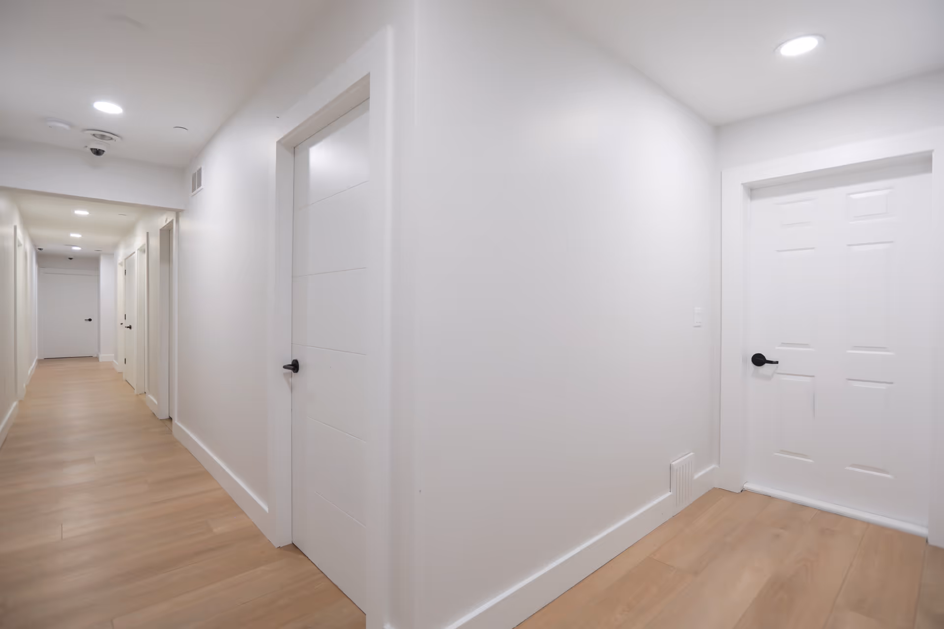 A clean, well-lit hallway with light wood flooring and white walls. Several white doors with black handles line the hallway, and recessed ceiling lights illuminate the space.