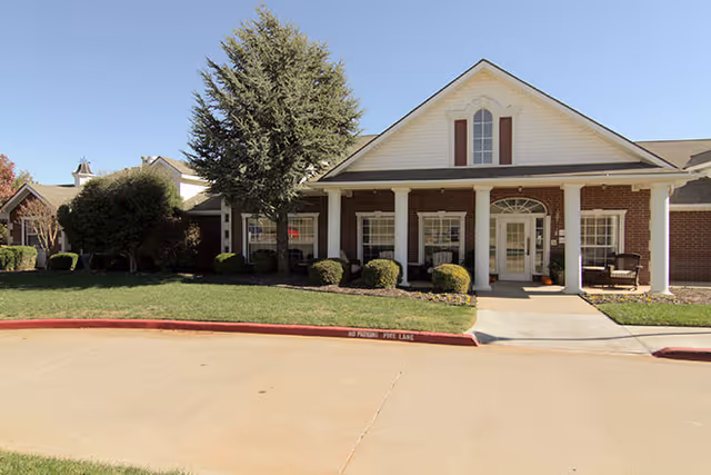 Front exterior of a brick building with white columns, a covered entrance, trimmed shrubs, and a driveway.