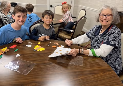 An elderly woman and two young boys sitting at a wooden table engaged in a craft activity with colorful paper cutouts shaped like leaves and flowers. In the background, other children and elderly people are also seated at tables in a well-lit room.
