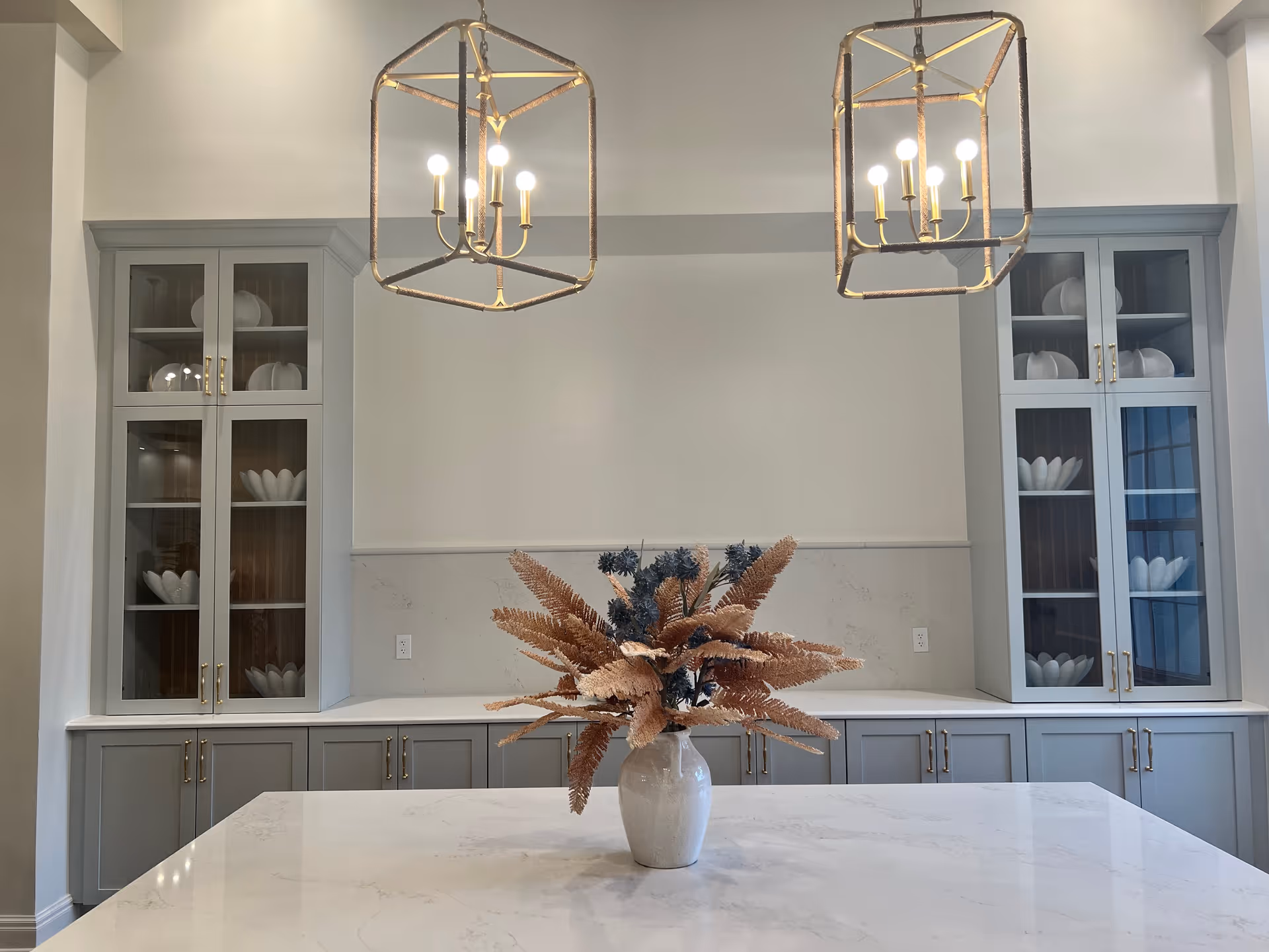 A modern kitchen interior featuring a large white marble island countertop with a vase of dried flowers in the center. Behind the island are gray cabinets with glass doors displaying white decorative dishes. Two geometric pendant lights with exposed bulbs hang from the ceiling above the island.