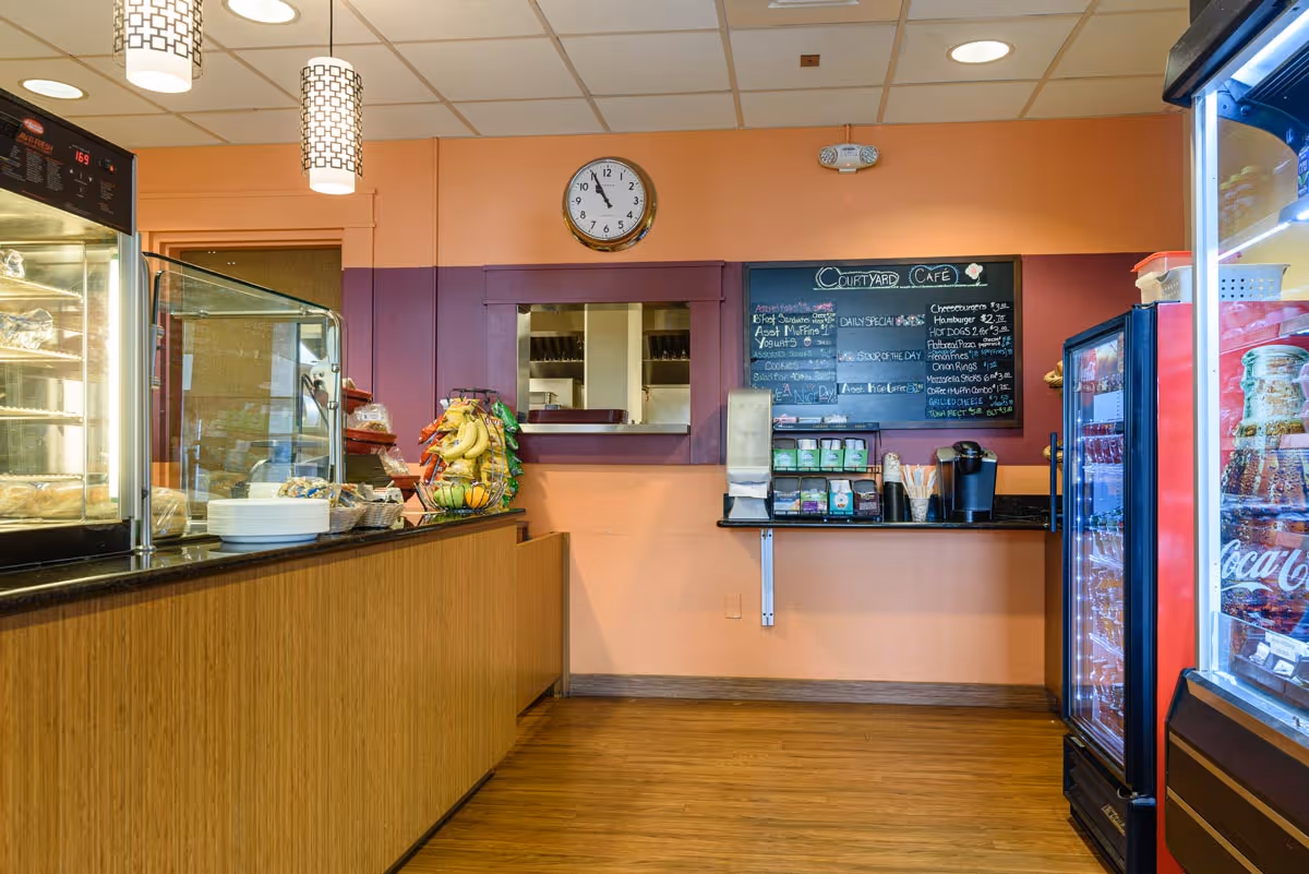 Interior view of a small café area with a wooden counter displaying plates, baskets of snacks, and a fruit stand. Behind the counter is a wall with a clock and a chalkboard menu labeled 'Courtyard Café'. To the right, there are refrigerated beverage coolers filled with drinks. The walls are painted peach and purple, and the floor is wooden.