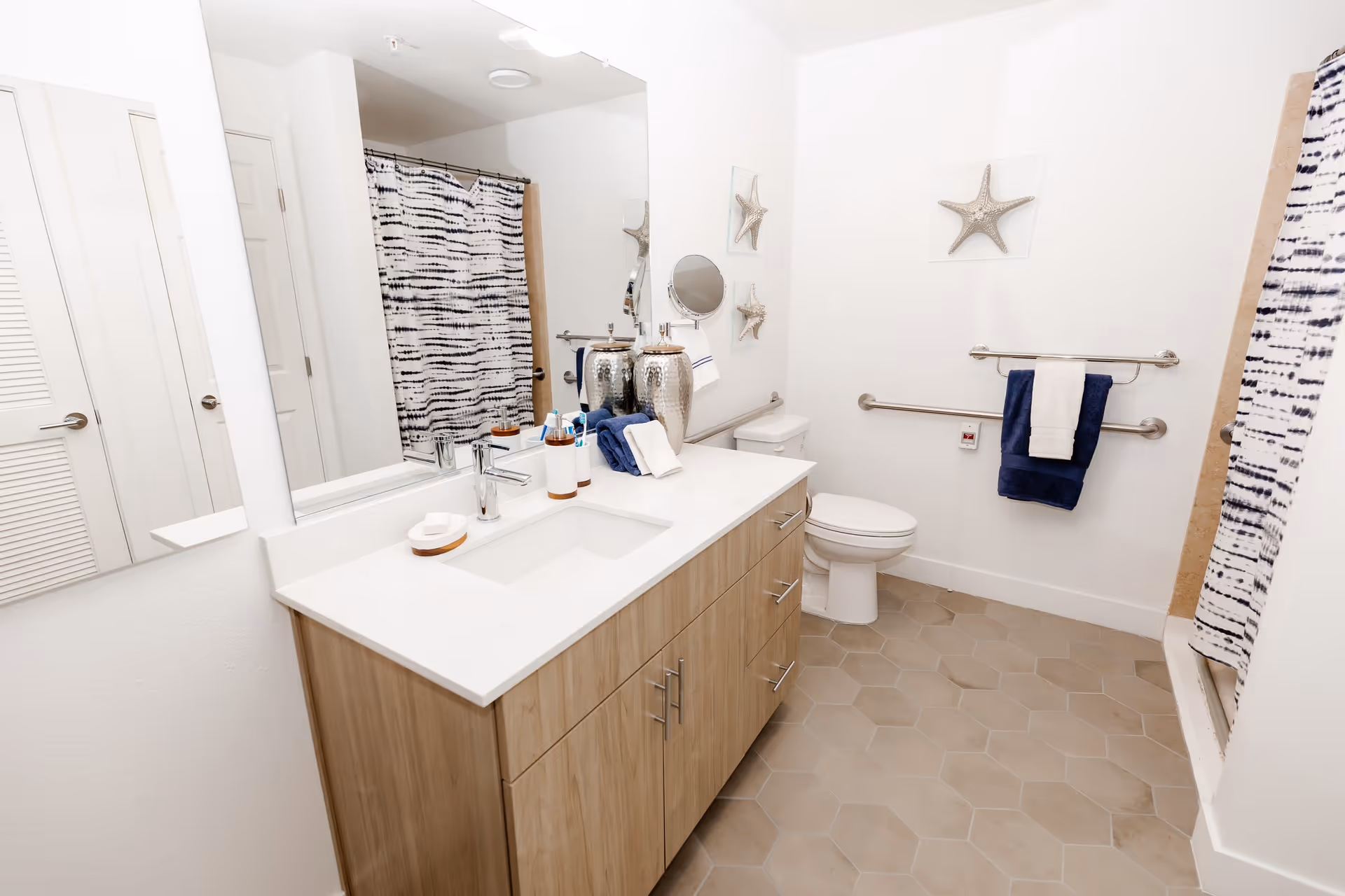 A clean and modern bathroom featuring a wooden vanity with a white countertop and built-in sink. Above the vanity is a large mirror. The bathroom includes a toilet with grab bars on the wall for accessibility. There is a shower with a black and white patterned curtain. The floor is covered with hexagonal beige tiles. Towels and decorative starfish wall art are also visible.