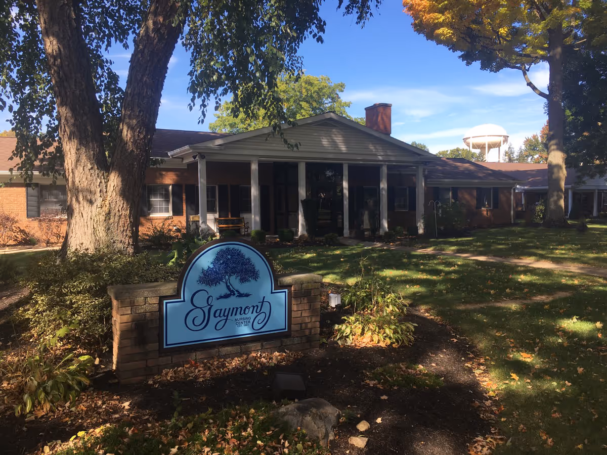 Exterior view of Gaymont Nursing Center, a single-story brick building with white columns at the entrance. There is a large tree and landscaped garden in front, along with a blue sign displaying the facility's name. A water tower is visible in the background under a clear blue sky.