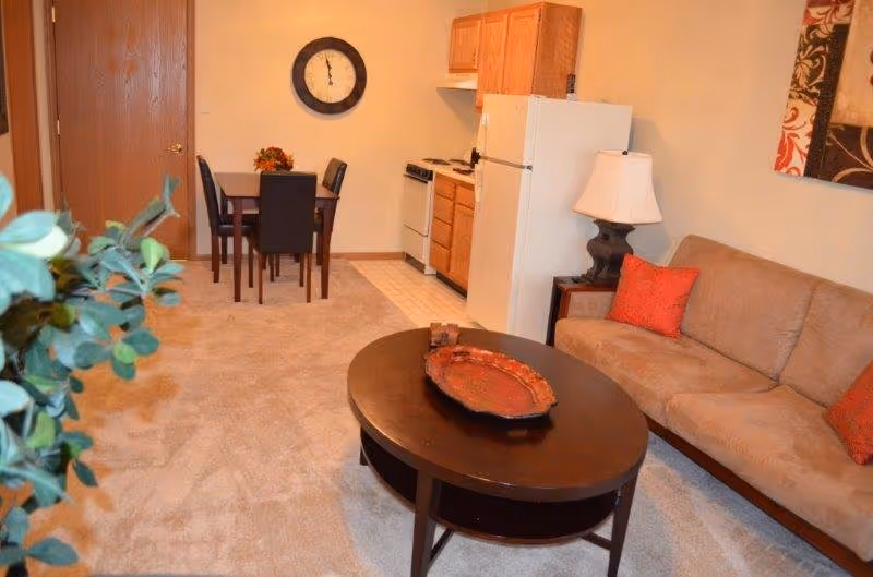 A cozy living area with a beige couch adorned with red pillows, a dark wooden oval coffee table with a decorative tray, a side table with a lamp, and a small dining table with four chairs near a kitchen area that includes a refrigerator, stove, and wooden cabinets. A large round wall clock is mounted on the wall above the dining table.