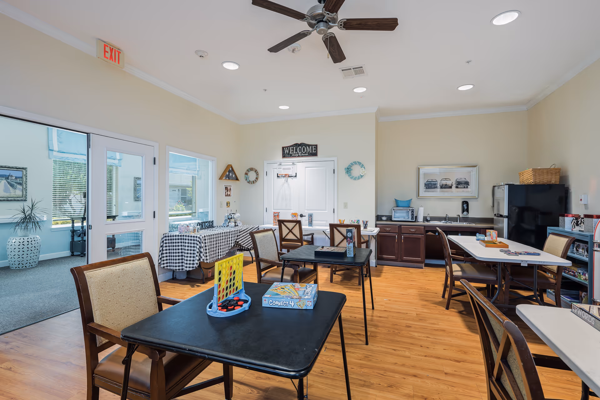 Bright multipurpose senior activity room with tables and chairs, board games on a table, a small kitchenette and a welcome sign on the wall.