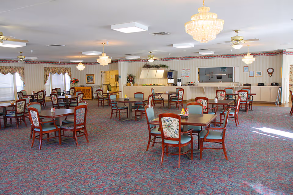 A spacious senior living dining room with multiple square tables and chairs arranged neatly on a patterned carpet. The chairs have floral upholstery on the backrests and teal seats. The room features chandeliers and ceiling fans, with a counter area in the back that includes an ice machine and various kitchen appliances. Windows with floral valances allow natural light into the room.