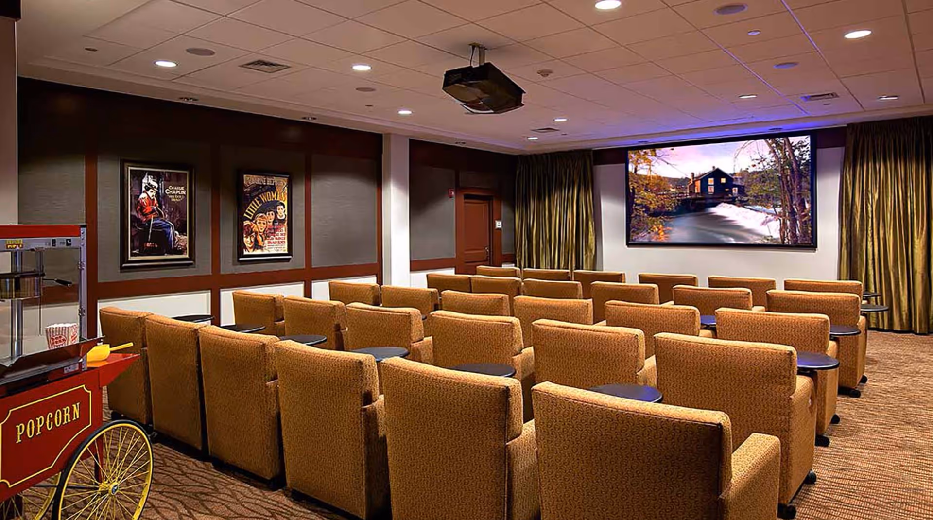 A cozy movie theater room with multiple rows of cushioned armchairs facing a large screen displaying a scenic image of a house by a river. The room has framed vintage movie posters on the wall, a popcorn machine on the left side, and ceiling-mounted projector. The room is warmly lit with recessed lighting and has carpeted floors.