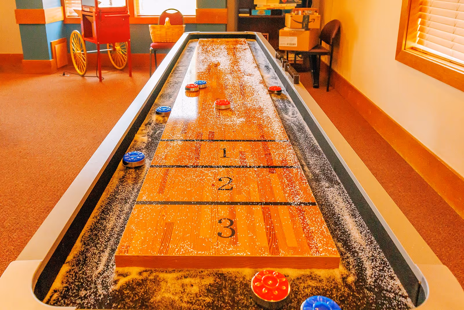 Indoor shuffleboard table with red and blue pucks scattered on the playing surface, surrounded by a carpeted room with windows, chairs, and a popcorn machine in the background.