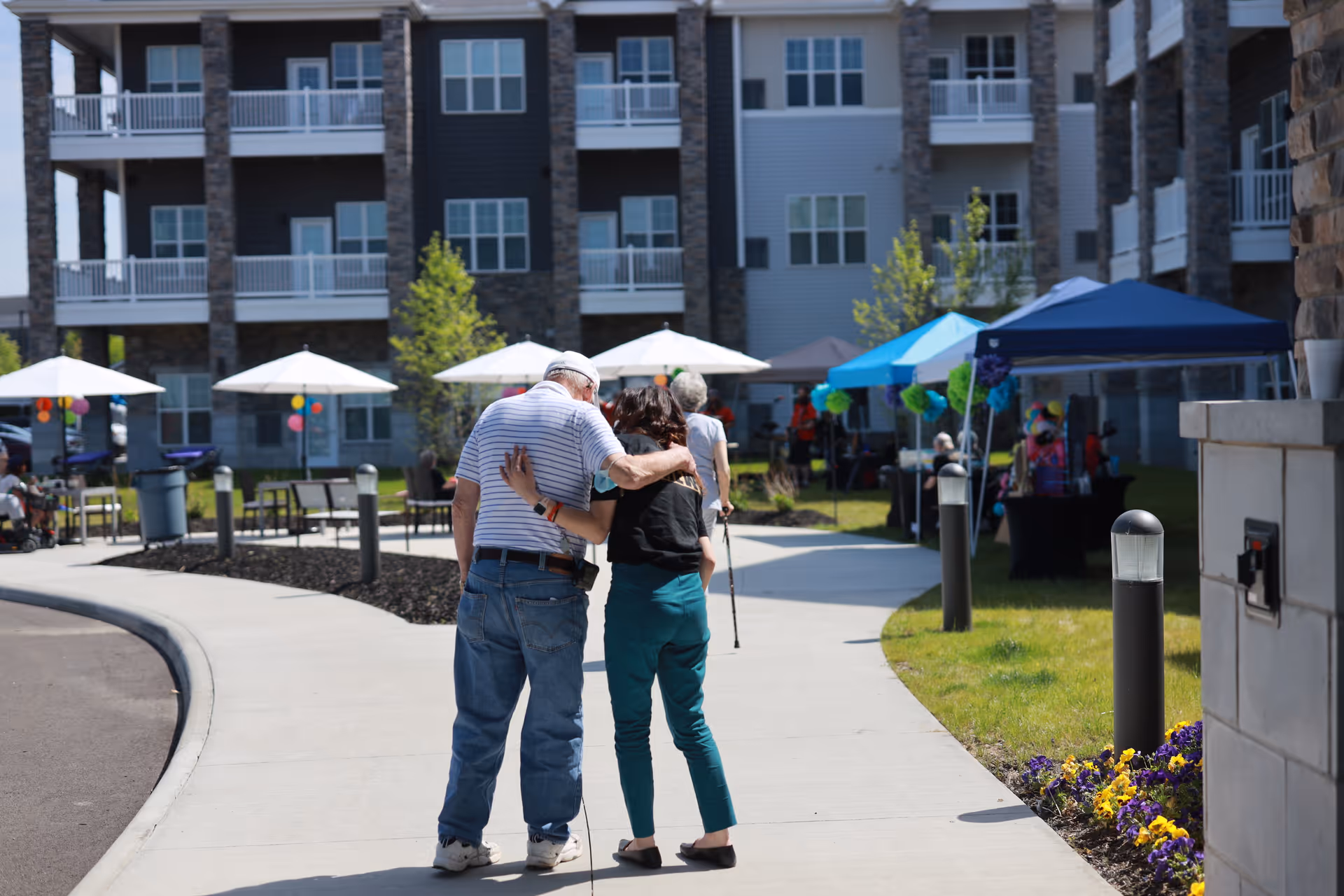 An elderly man and a woman walking arm in arm on a paved pathway outside a multi-story residential building with balconies. There are white umbrellas, tents, and people in the background, suggesting an outdoor event or gathering at the community.