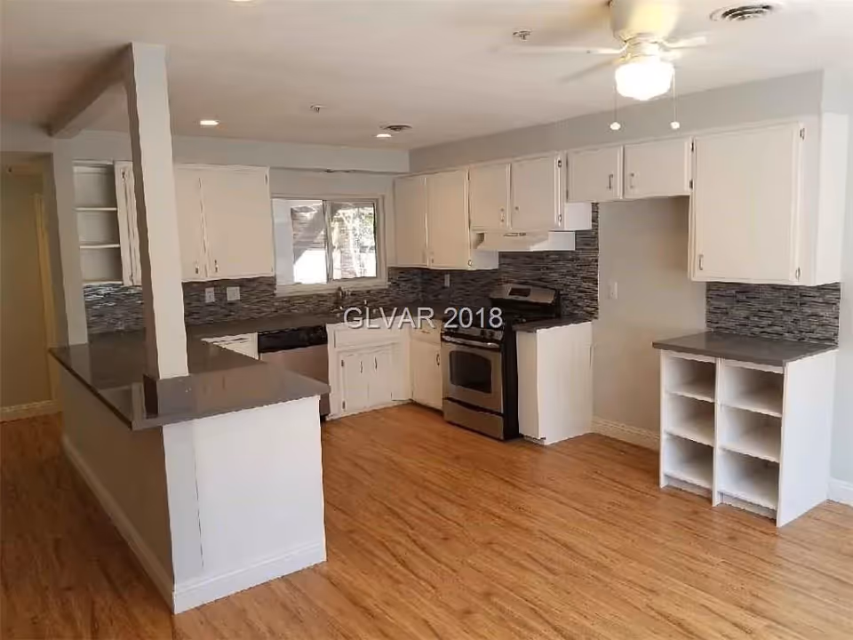 A modern kitchen with white cabinets, gray countertops, a stainless steel stove, and a ceiling fan with a light. The kitchen has a wooden floor and a window above the sink. There is a tiled backsplash in shades of gray and black.