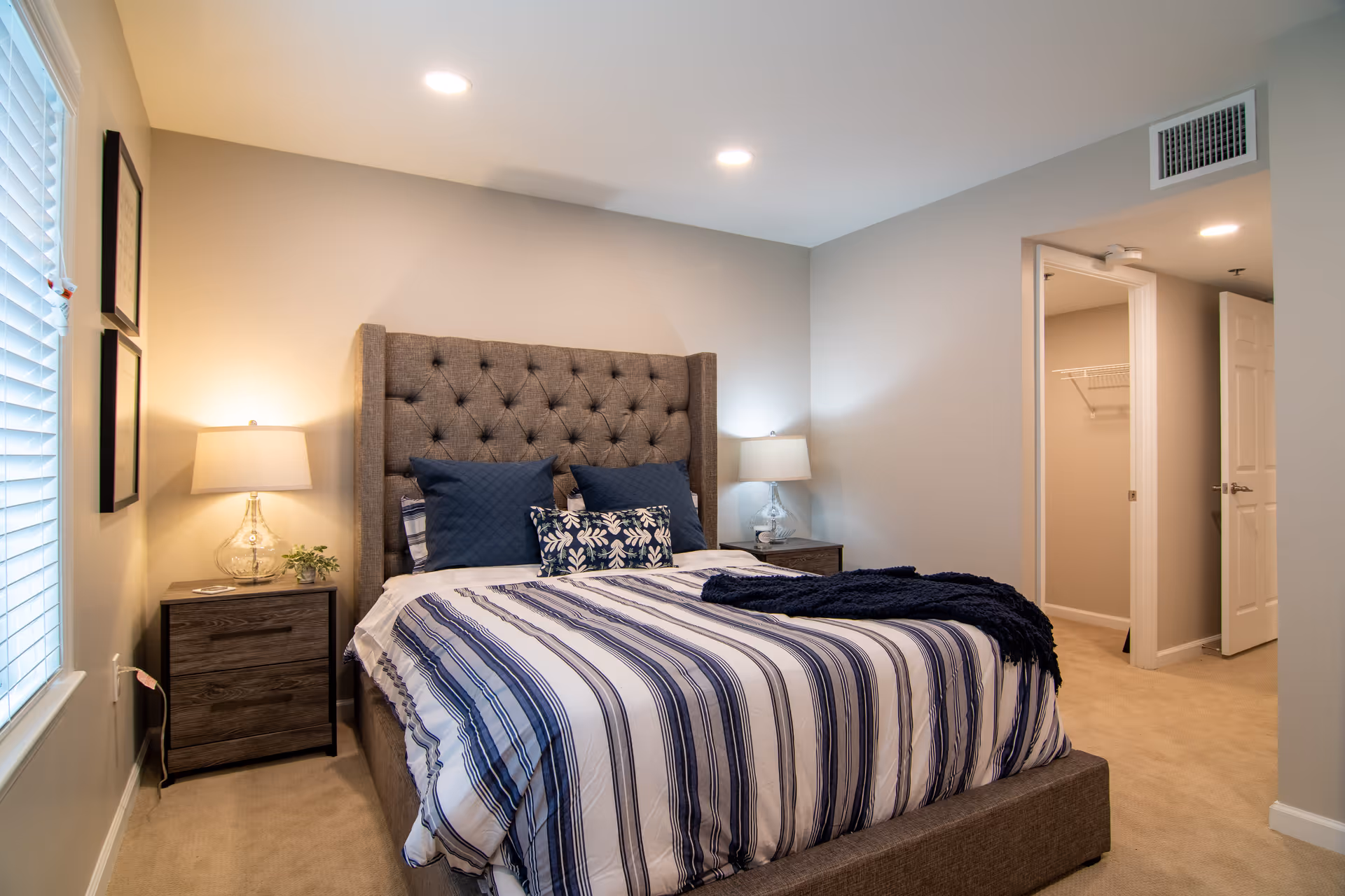 A neatly made bed with a tufted headboard in a bedroom. The bed has striped bedding in shades of blue and white, with several pillows including a decorative one. There are two bedside tables, each with a lamp and one with a small plant. The room has beige walls, carpeted floor, and a window with blinds on the left side. An open door reveals a walk-in closet with shelving inside.