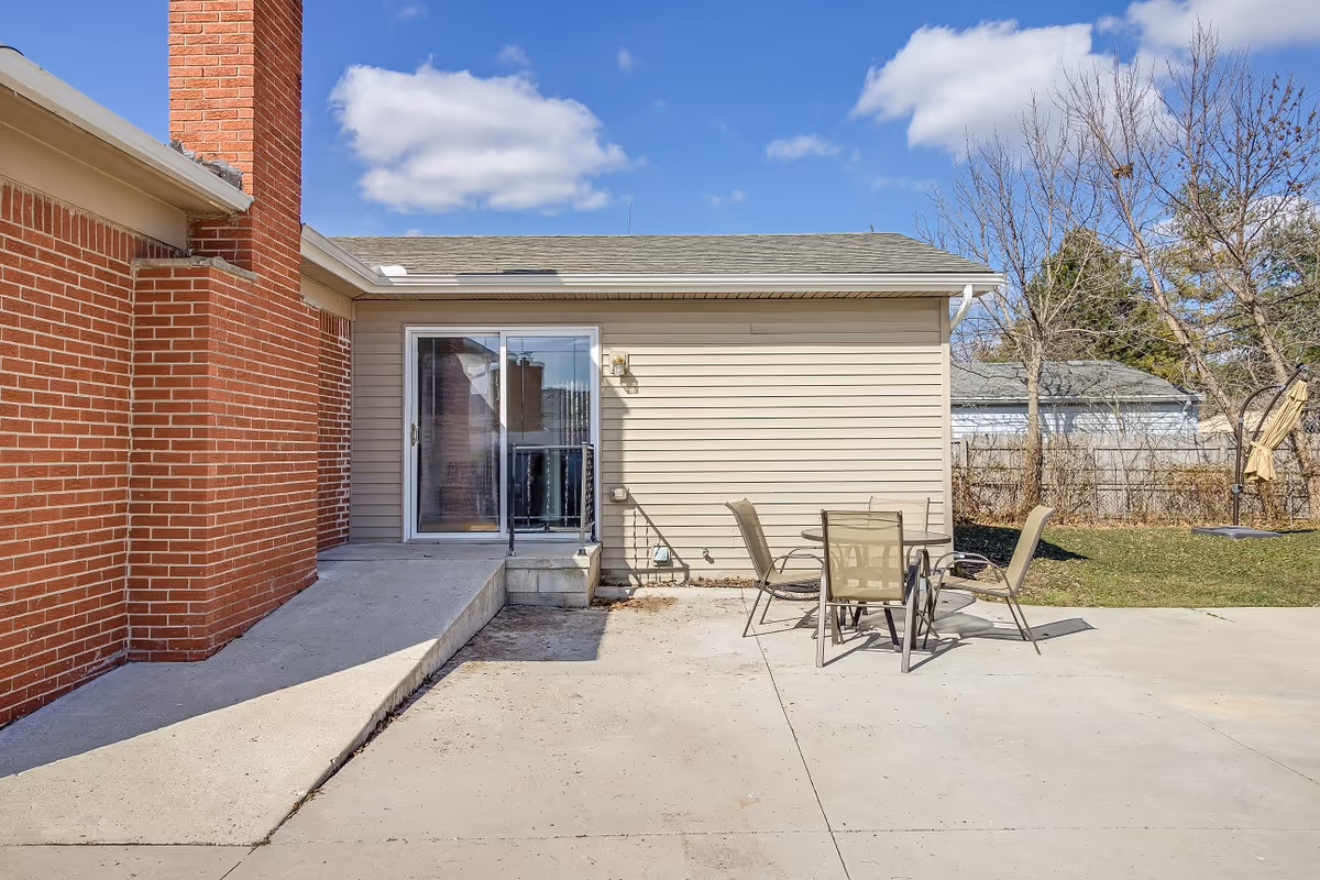 Concrete patio with a sliding glass door, brick chimney, ramp, and a table with chairs beside a beige-sided building under a blue sky.