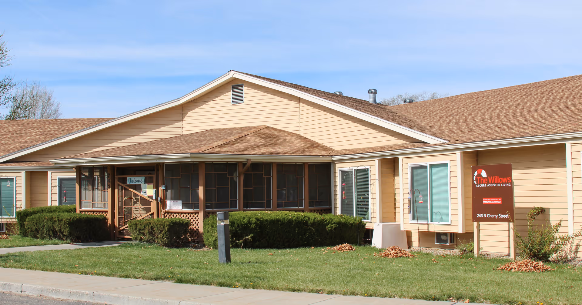 Exterior view of a single-story assisted living facility building with beige siding and a brown shingled roof. There is a screened porch area with a 'Welcome' sign, green bushes in front, and a sign on the lawn that reads 'The Willows Secure Assisted Living' with an address below. The sky is clear and blue.