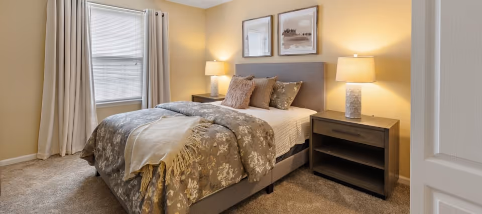 A cozy bedroom with a neatly made bed featuring floral patterned bedding and multiple pillows. There are two wooden nightstands on either side of the bed, each with a lamp. A window with closed blinds and beige curtains is on the left wall. Two framed abstract artworks hang above the bed. The walls are painted a warm beige color and the floor is carpeted.