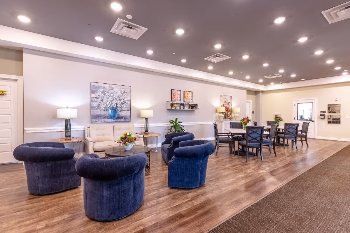 A spacious and well-lit common area in a senior living facility with several blue upholstered armchairs arranged around a round coffee table, a beige sofa against the wall, and multiple dining tables with chairs. The walls are decorated with floral artwork and lamps on side tables, and the floor is wooden with a carpeted section.