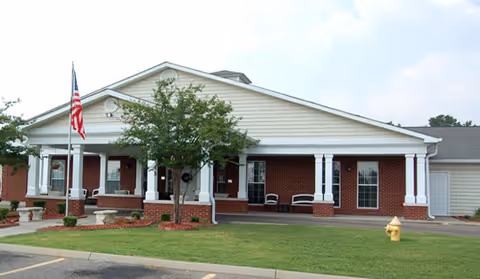 Front exterior view of a single-story brick building with white columns and a covered porch. There is an American flag on a flagpole in front, a small tree, and a yellow fire hydrant on the lawn.