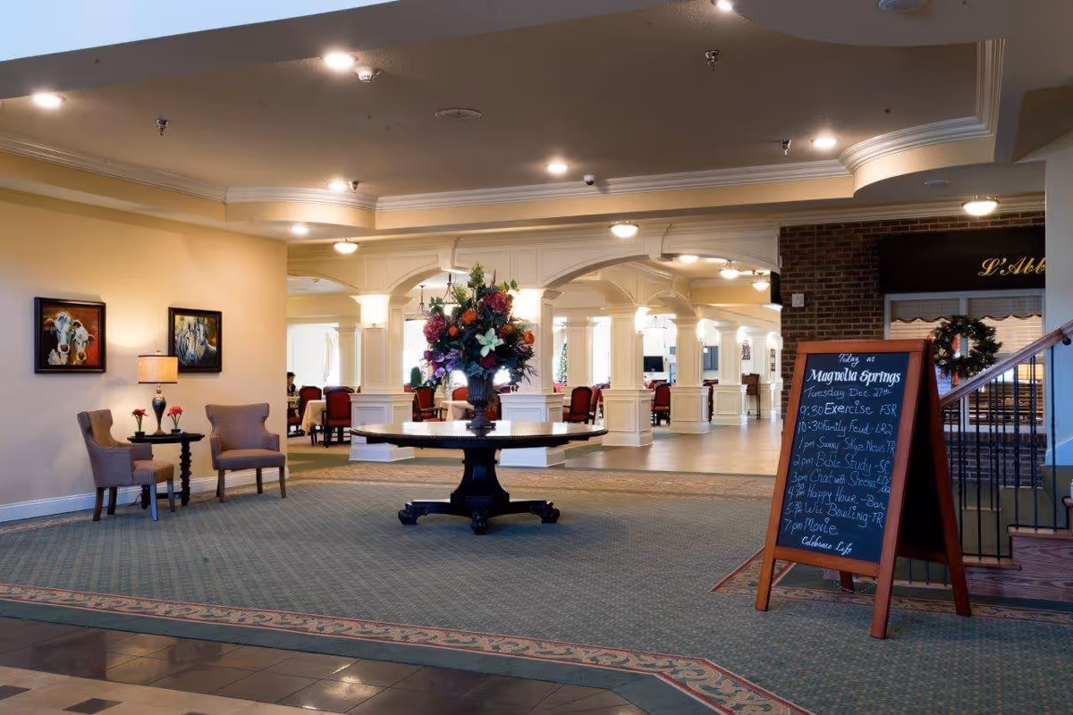 Interior view of a senior living facility lobby area with a round table holding a large floral arrangement in the center. To the left, there are two chairs with a small table and lamp between them, and two paintings on the wall. In the background, there is a dining area with red chairs and white columns. On the right side, a chalkboard sign lists daily activities and a staircase is partially visible.