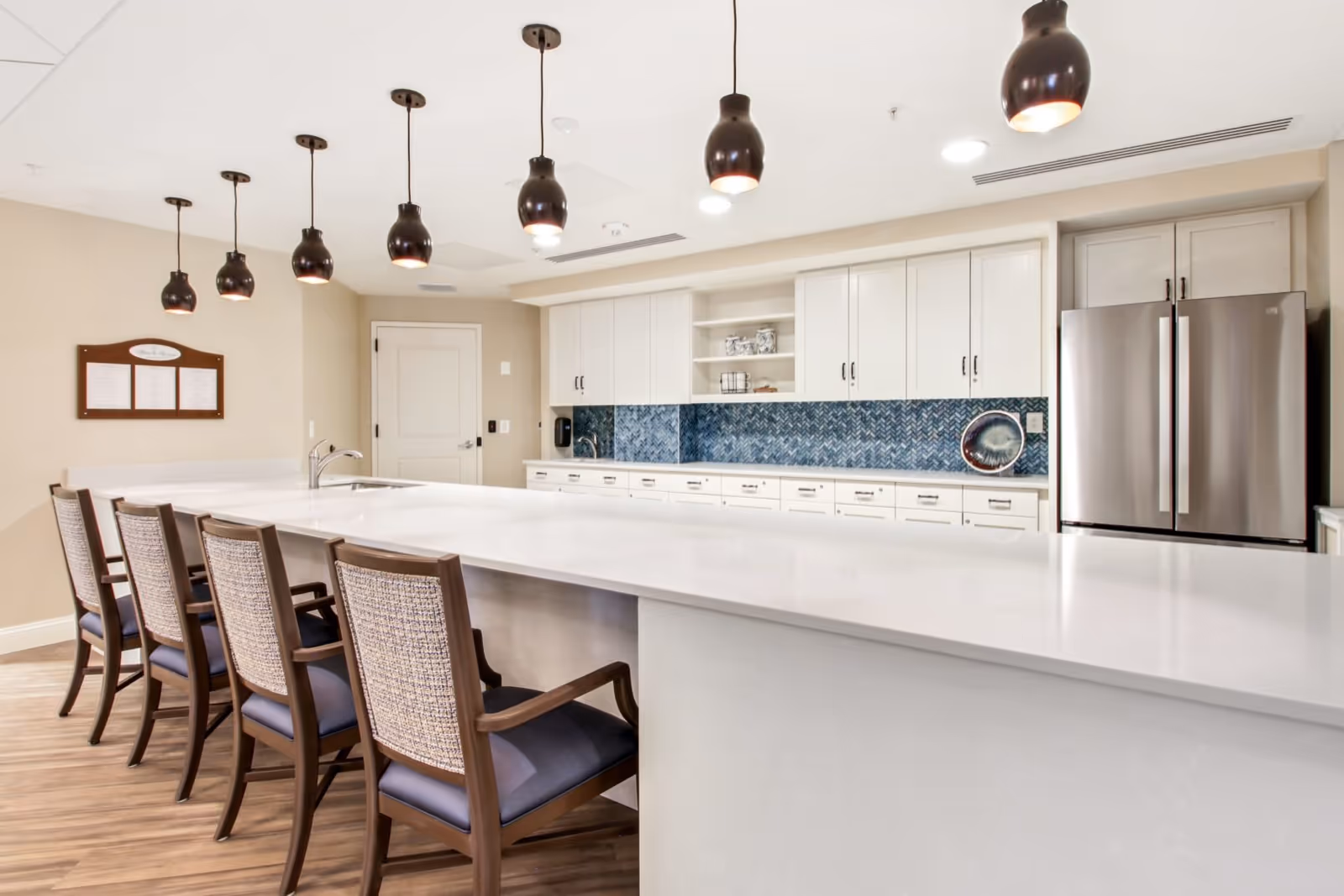 Bright communal kitchen featuring a long white island with five chairs, pendant lights, white cabinetry, blue tile backsplash, and a stainless steel refrigerator.