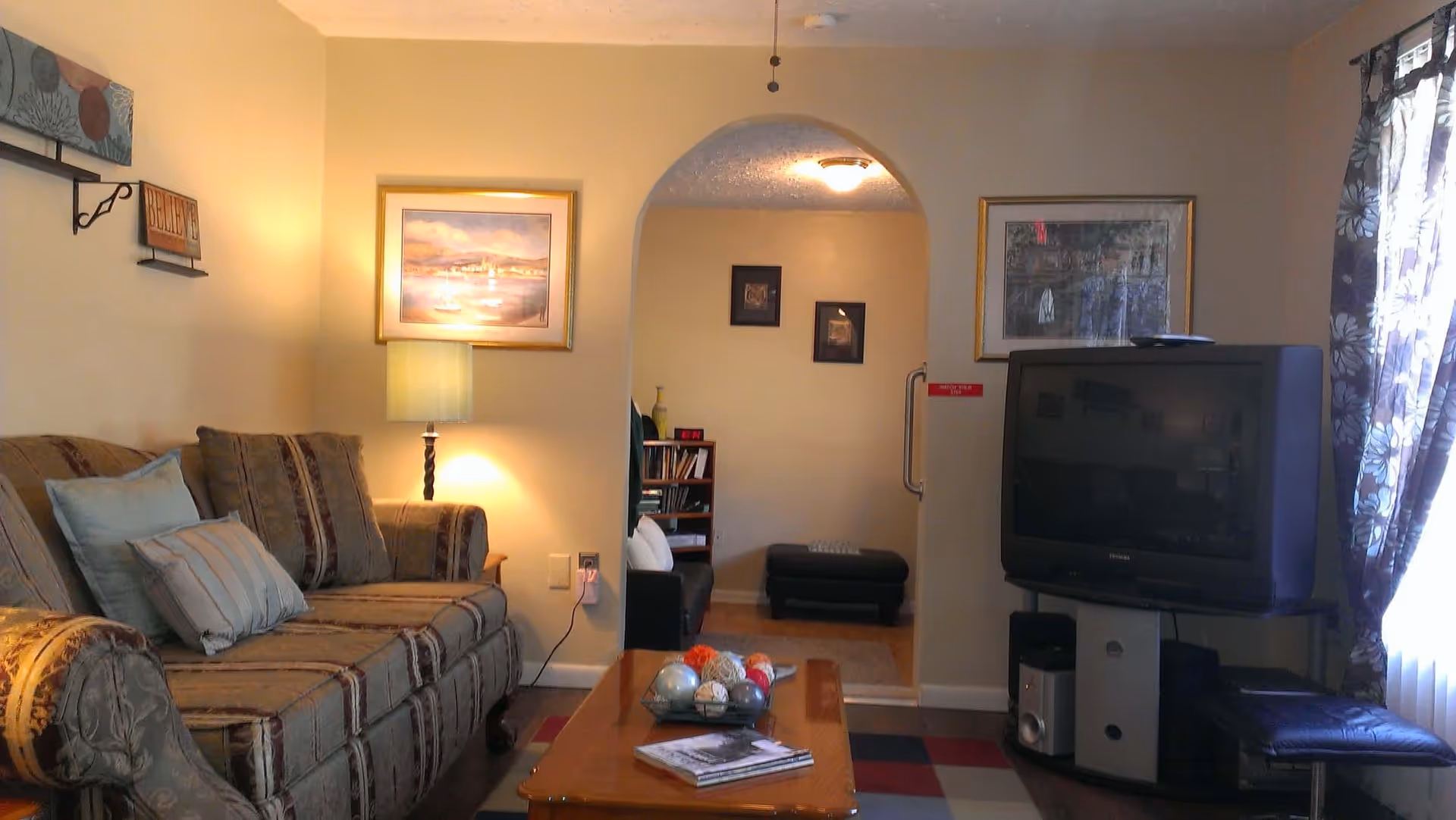 A cozy living room with a patterned sofa adorned with cushions on the left, a wooden coffee table with decorative balls and magazines in the center, and a large CRT television on a stand to the right. The room has beige walls with framed artwork, a floor lamp beside the sofa, and an arched doorway leading to another room with a bookshelf and additional seating. A window with floral curtains allows natural light to enter from the right side.