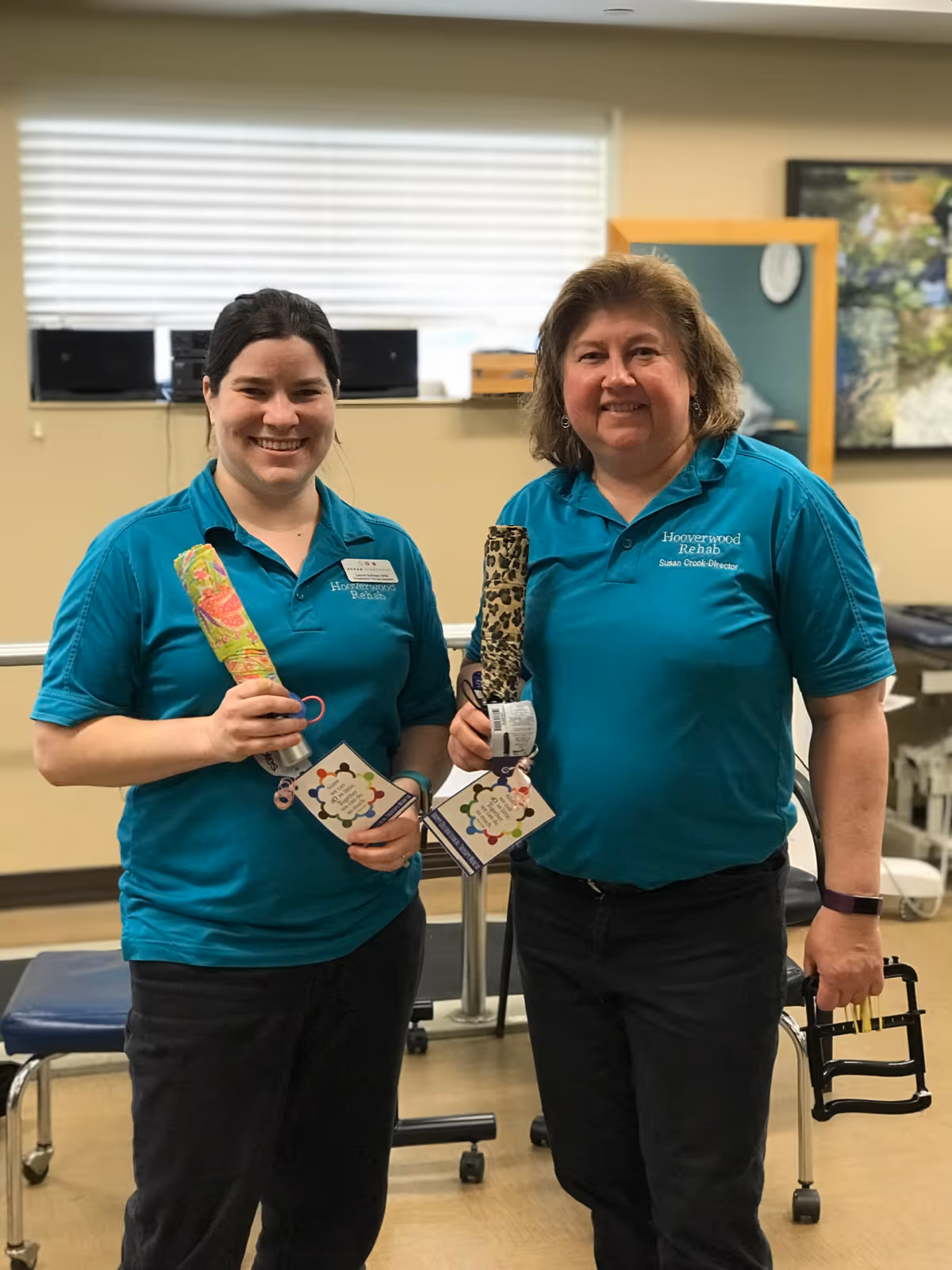Two women standing indoors wearing teal polo shirts with Hooverwood Rehab logos, each holding a folded umbrella and smiling at the camera. The background shows a window with blinds, a mirror, and some furniture.