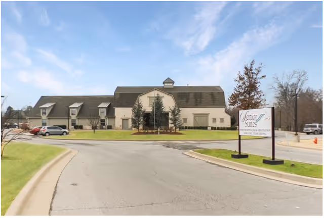 Exterior view of a large building with a sloped roof and multiple windows, surrounded by a parking lot and grassy areas under a partly cloudy sky. A sign in front reads 'Senior Suites' with additional text about private suites, rehabilitation, and assisted living.