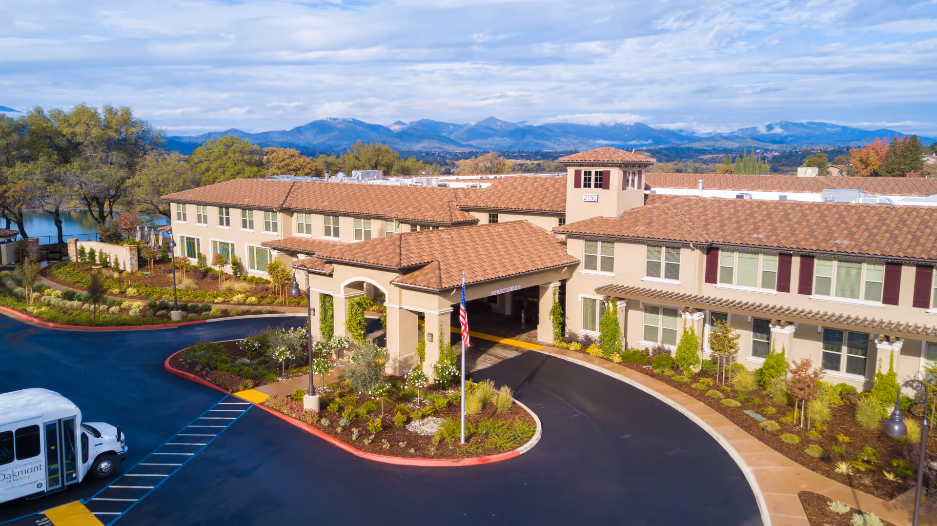 Aerial view of Oakmont of Redding senior living facility showing a large building with beige walls and a red-tiled roof. The entrance has a covered driveway with an American flag in front. Surrounding the building are landscaped gardens, trees, and a parking area with a white shuttle bus. In the background, there are mountains under a partly cloudy sky.