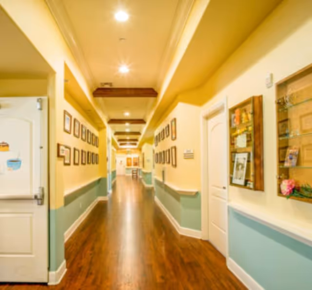 A long, well-lit hallway in a senior living facility with wooden flooring, light yellow walls with a teal lower half, and framed pictures hanging on both sides. The ceiling has recessed lighting and wooden beams. There are doors and display cases along the hallway.
