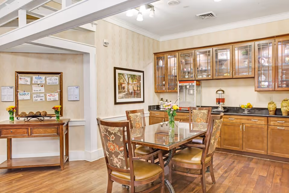 Bright dining area with a wooden table and upholstered chairs, glass-front cabinets and a bulletin board on the wall.