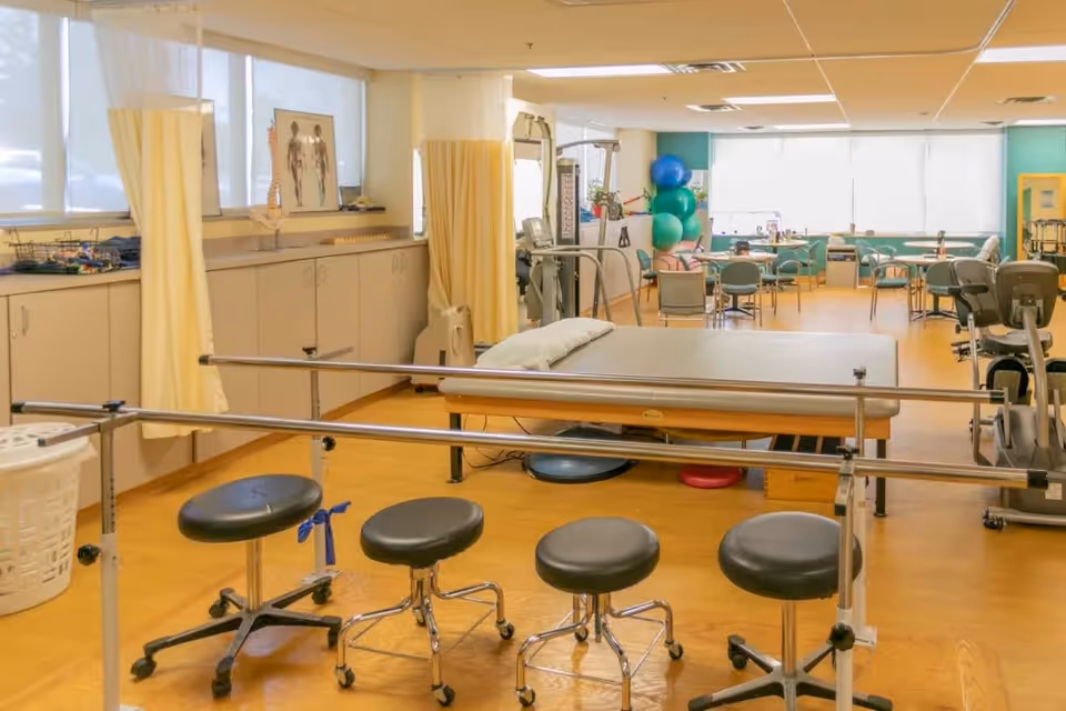 A bright rehabilitation/therapy room with parallel bars, a treatment table, rolling stools, exercise equipment and a seating area in the background.
