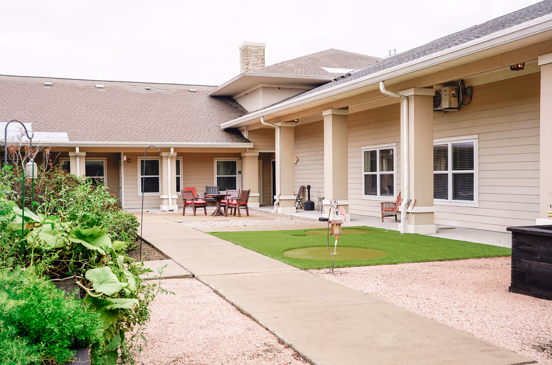 Outdoor courtyard area of Orchard Park at Victory Lakes Assisted & Senior Living featuring a concrete walkway, green plants on the left, a small putting green, and several chairs and tables arranged near the building with beige siding and white-trimmed windows.