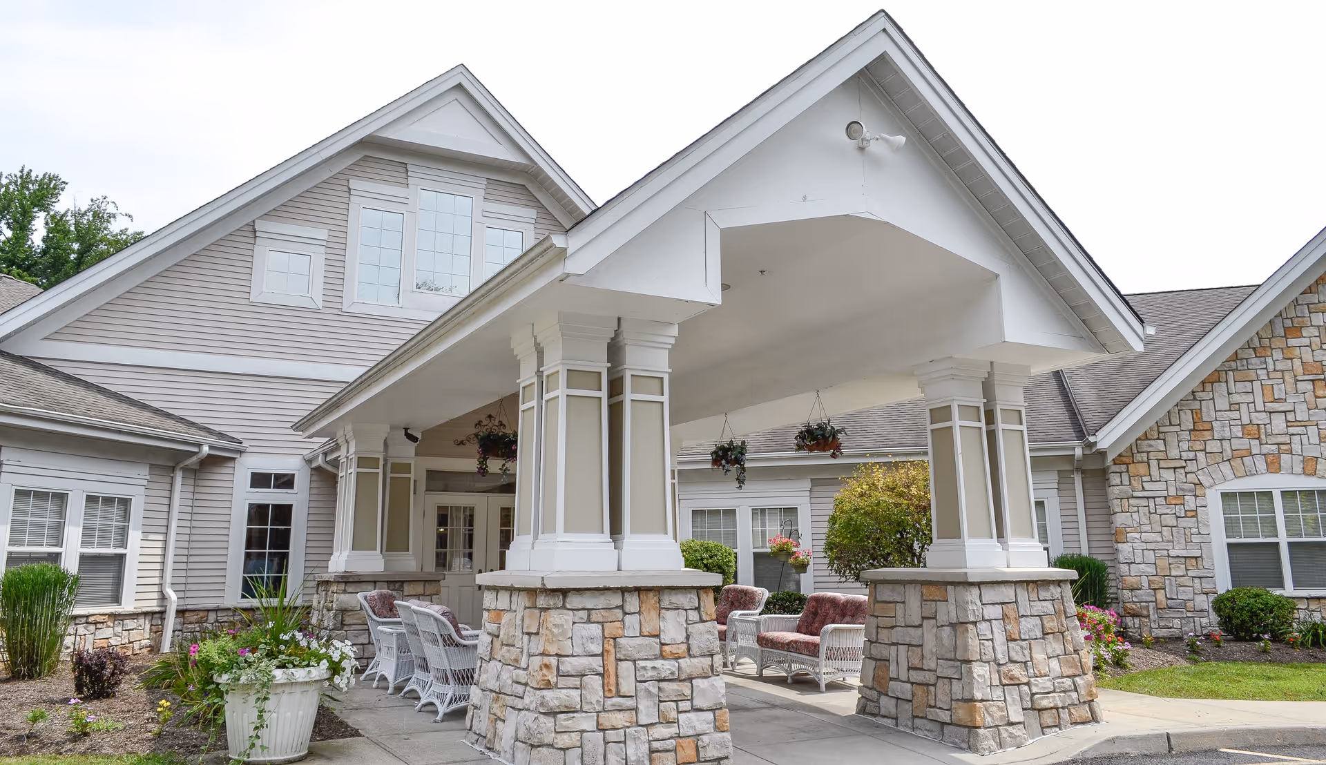 Covered entrance/porte-cochere of a beige stone-and-siding senior living building with outdoor wicker seating and hanging plants.