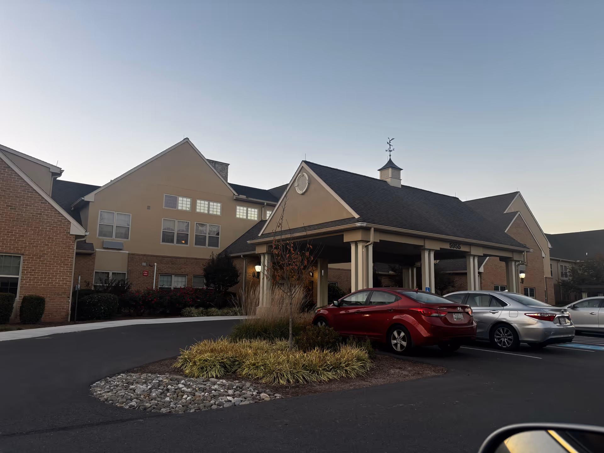 Exterior view of a retirement community building with a covered entrance and several parked cars in front. The building has a combination of brick and beige siding with multiple windows and a weather vane on top of the entrance roof. Landscaping with bushes and ornamental grasses surrounds the driveway.