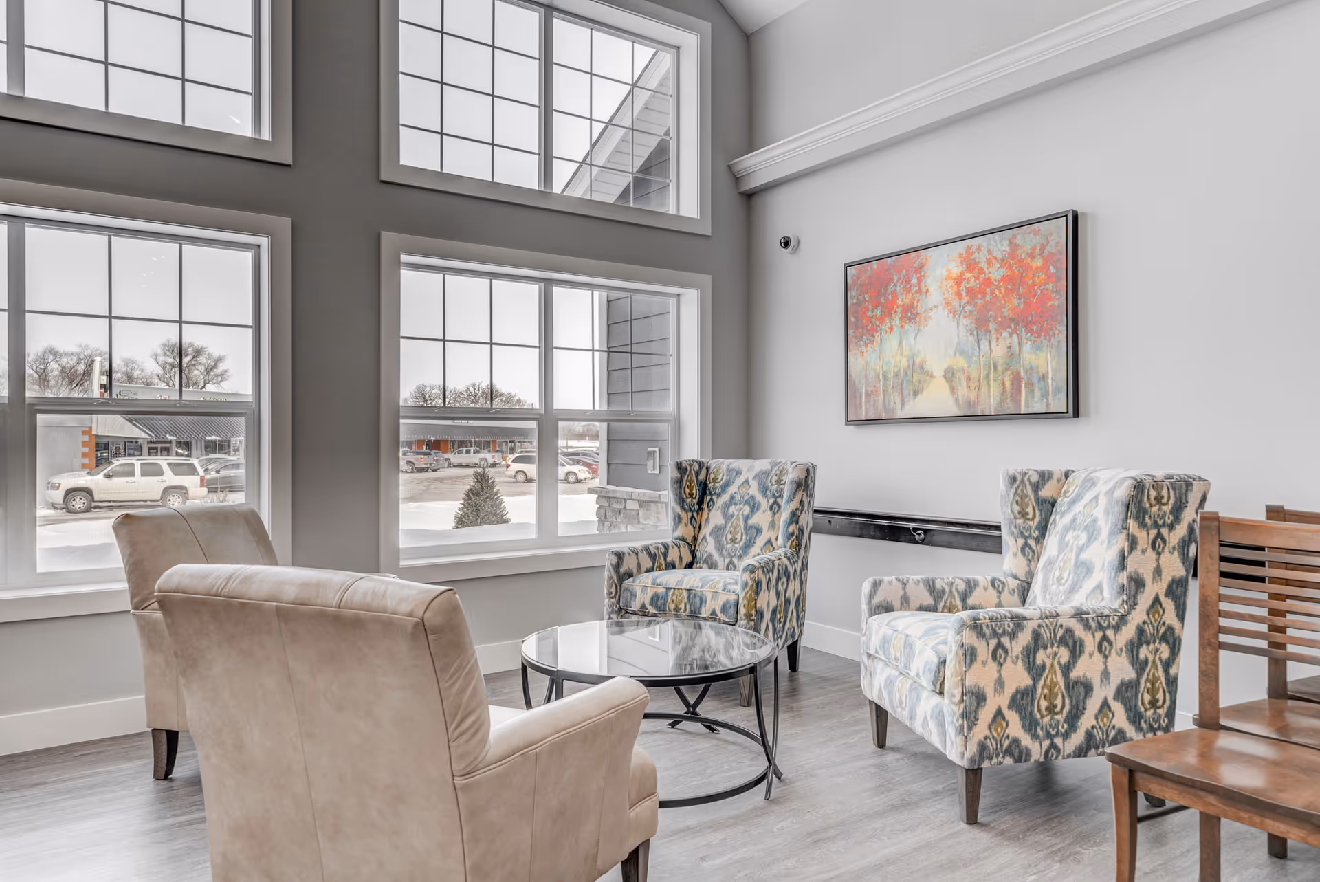 Bright sitting area with upholstered armchairs, a glass coffee table, large windows and framed artwork on the wall.