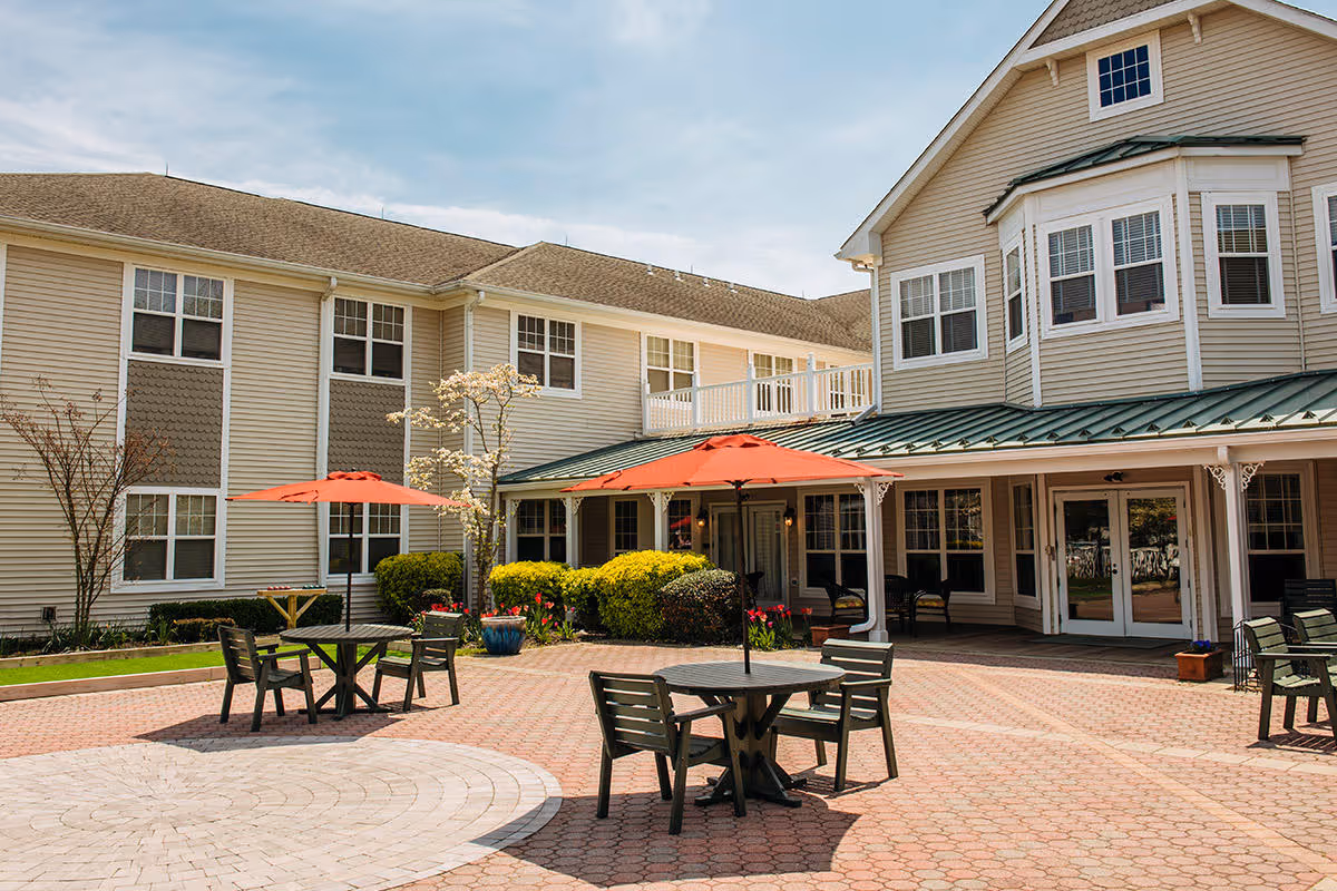 Outdoor courtyard area of a senior living facility with several tables and chairs under red umbrellas, surrounded by a two-story beige building with white trim and multiple windows. There are some bushes, small trees, and potted plants around the courtyard.