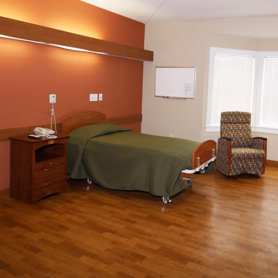 A senior living bedroom with a single bed on casters, a wooden nightstand, and an upholstered armchair by windows.