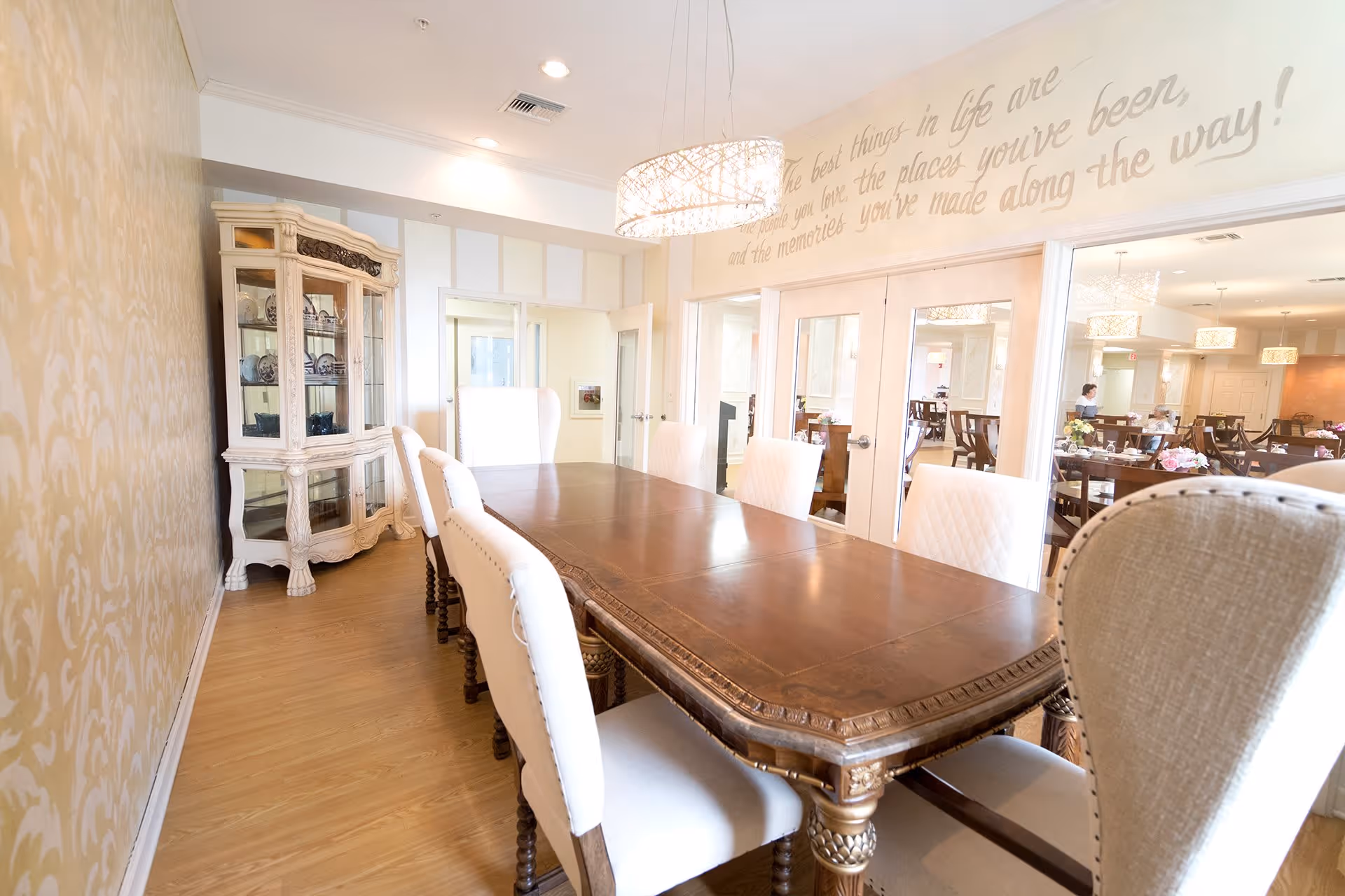 A bright dining room with a long wooden table surrounded by white upholstered chairs. A decorative white cabinet with glass doors is placed against the wall, and a modern chandelier hangs above the table. The walls are light-colored with a subtle pattern, and there is a quote written on the wall above the glass doors that lead to another dining area with more tables and chairs.
