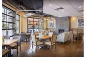 A spacious and modern dining area with multiple tables and chairs arranged neatly. Large windows allow natural light to fill the room. There is a fireplace on one wall with framed artwork above it. The ceiling has exposed ductwork and contemporary lighting fixtures.