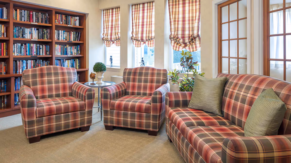 A cozy living room area with plaid upholstered furniture including a sofa and two armchairs. The room features a bookshelf filled with books, three windows with plaid Roman shades, a small round side table with decorative items, and some indoor plants near the windows.