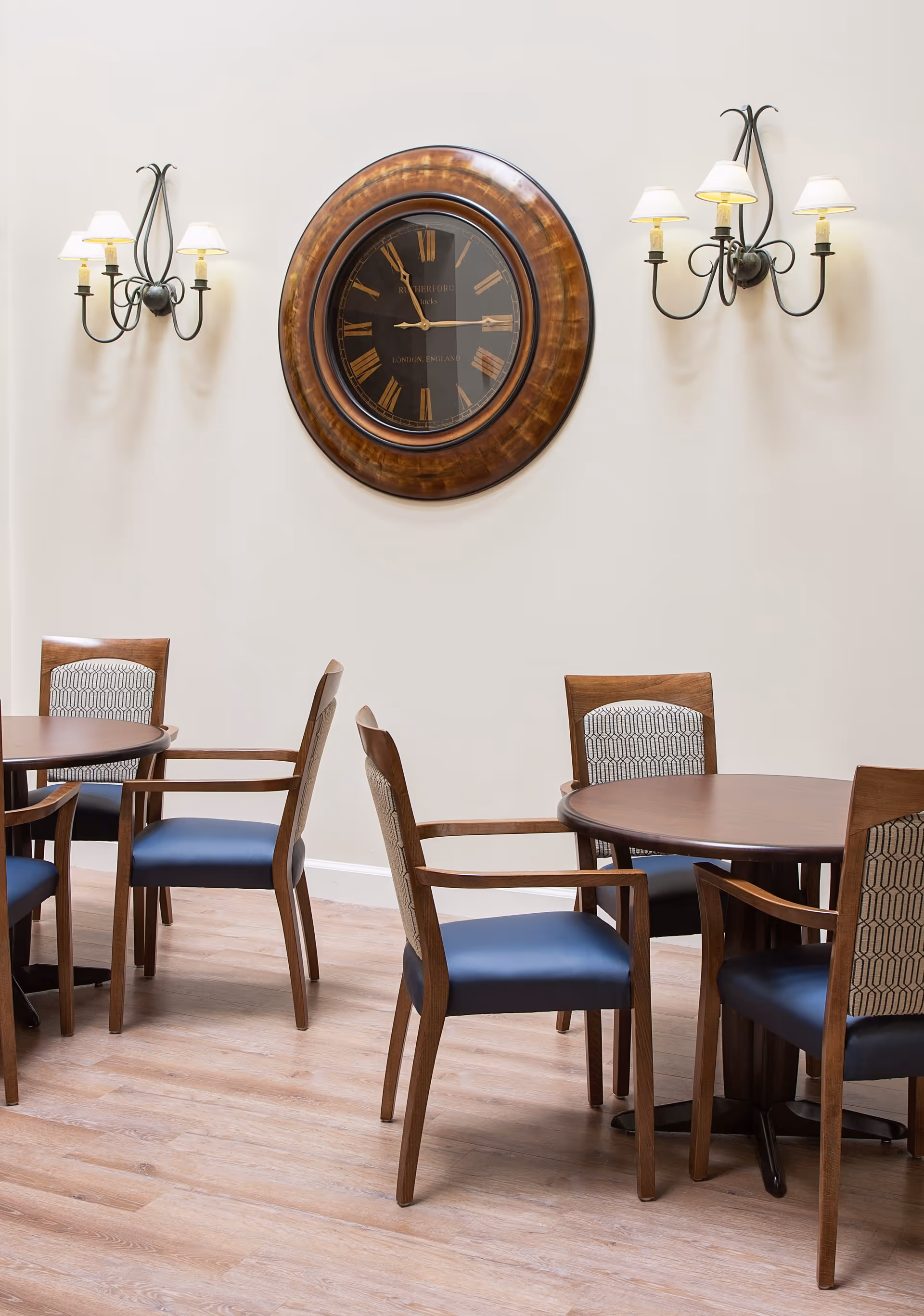 Interior view of a dining area with wooden round tables and chairs with blue cushions and patterned backs. A large round wall clock with Roman numerals is mounted on a white wall, flanked by two black metal wall sconces with three lamps each. The floor is light wood.