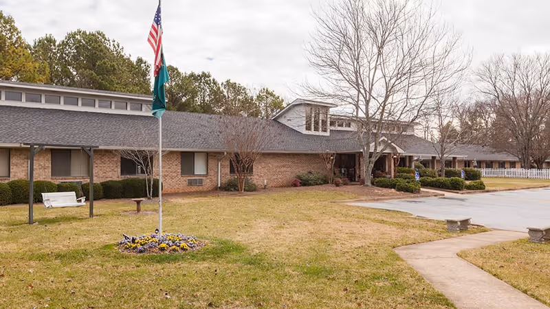 Exterior view of a single-story brick assisted living facility with a gray shingled roof, a flagpole with American and state flags, a white swing bench, and a small flower bed in the front lawn. There are leafless trees and a paved parking area with handicap parking signs.