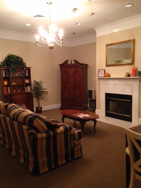 A cozy living room with a striped sofa facing a wooden coffee table and a white fireplace with a gold-framed mirror above it. There is a wooden cabinet and bookshelf with decorative items and a potted plant in the corner. A chandelier hangs from the ceiling providing warm lighting.