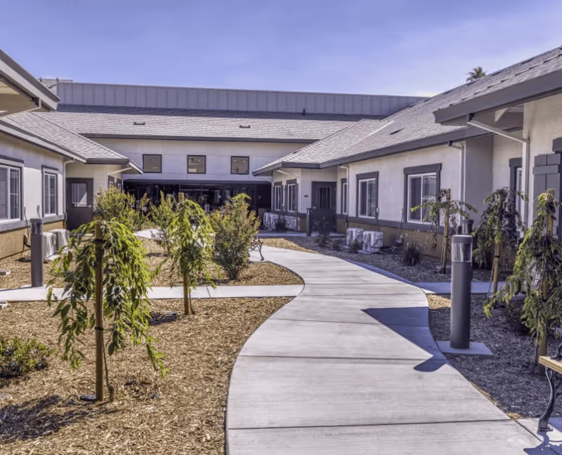 Curved concrete pathway leading through a landscaped outdoor courtyard with small trees and shrubs, surrounded by single-story buildings with windows and gray roofs under a clear blue sky.
