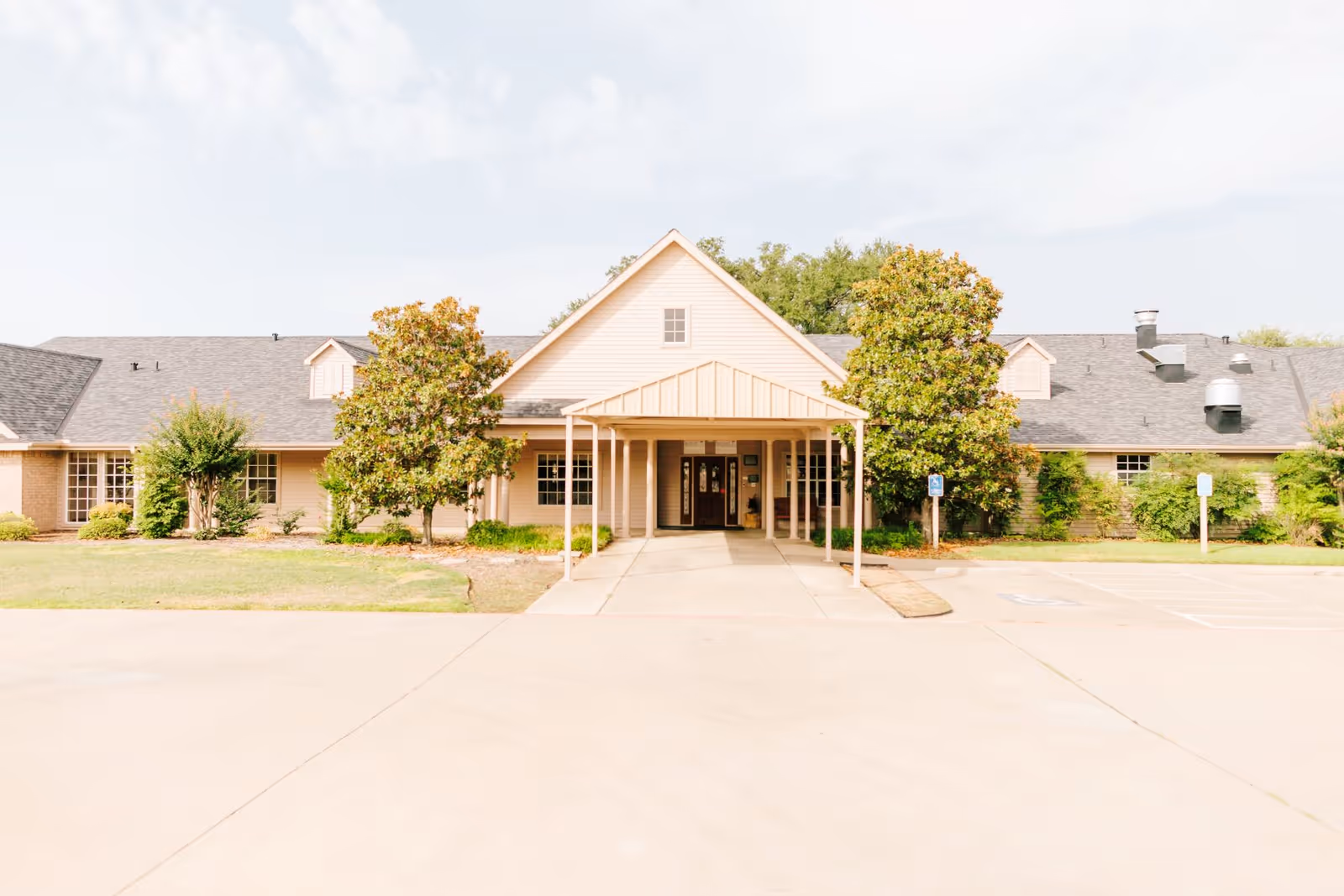 Front entrance of a single-story senior living building with a covered porte-cochère, trees, and a parking area.