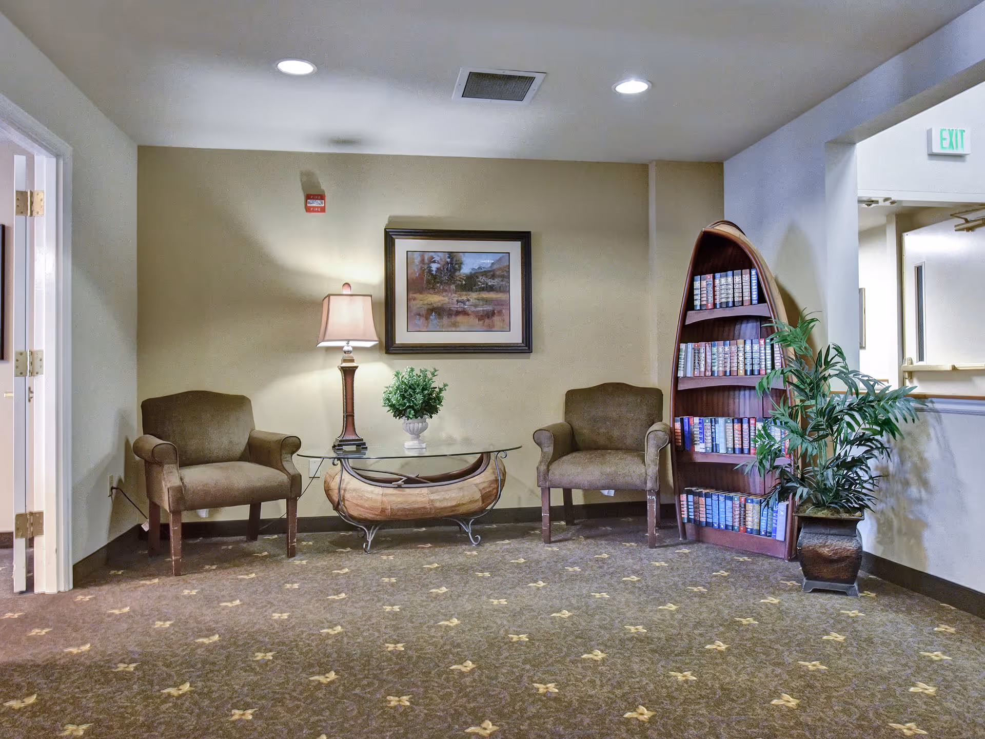 A cozy seating area in a senior living facility with two brown upholstered armchairs, a glass-top coffee table with a wooden base, a table lamp, a small potted plant, a framed landscape painting on the wall, a boat-shaped bookshelf filled with books, and a potted plant on the floor. The area is carpeted and well-lit with recessed ceiling lights.