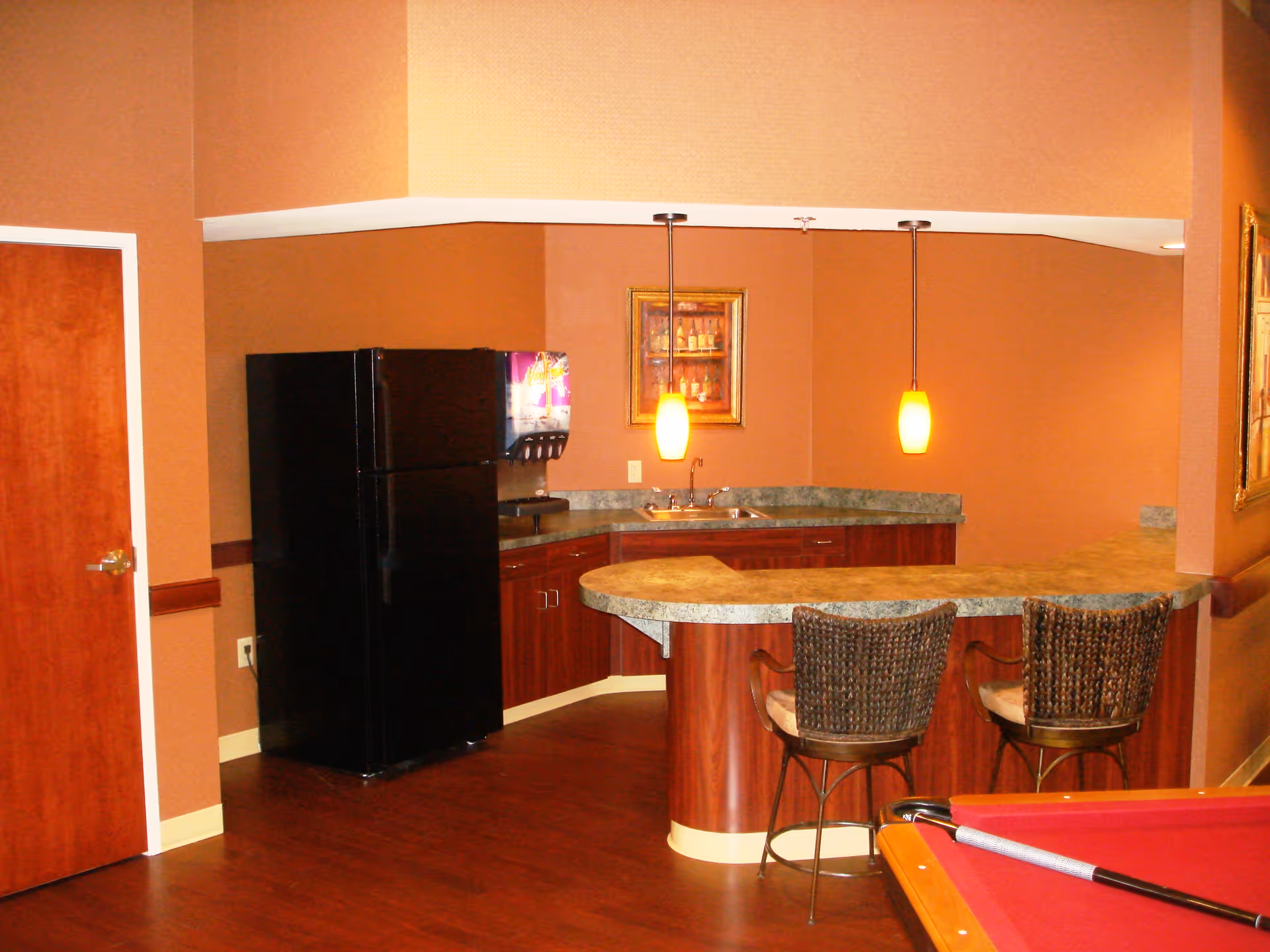 Small kitchenette and bar area with a curved granite countertop, two wicker bar stools, a black refrigerator, pendant lights, and a nearby pool table corner.