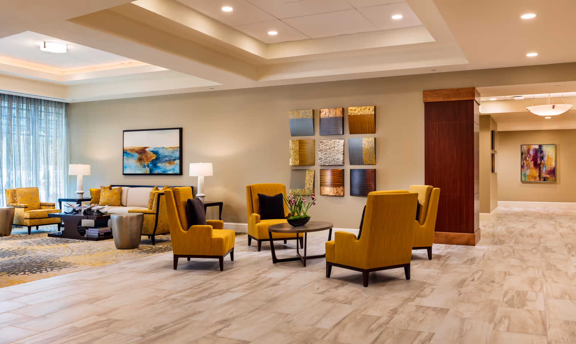 A spacious and well-lit living room area in a senior living facility with mustard yellow armchairs arranged around a round wooden table with a flower arrangement. In the background, there is a beige sofa with yellow cushions, two white table lamps, and a coffee table with decorative items. The walls are adorned with modern abstract artwork, and large windows with sheer blue curtains allow natural light to fill the room. The floor is covered with light-colored tiles and a patterned area rug.
